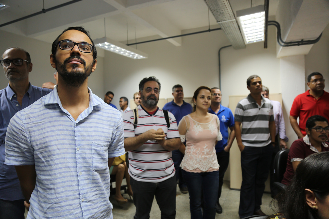 parte do publico que participou da cerimônia, em primeiro plano um homem jovem, de camisa padrão xadrez na cor azul; atrás e ao lado dele, dois homens com camisetas de listras horizontais e uma mulher com uma blusa em tons claros de rosa