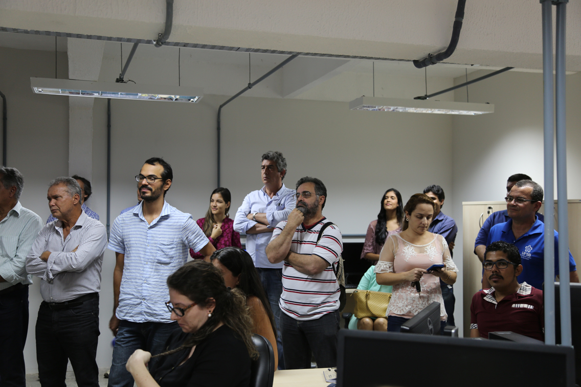 parte do público participante da solenidade; há mulheres sentadas ao redor de uma mesa e homens e mulheres em pé, um pouco atrás